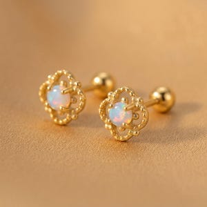 May include: A pair of gold stud earrings featuring a central round iridescent opal stone set within a decorative four-petal floral frame. Each earring includes a smooth gold post and a spherical back closure, showcasing a polished metallic finish.