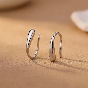 Puede incluir: Un par de pendientes tipo ear cuff de tono plateado con un diseño liso, pulido y en forma de gota. Cada pieza tiene una estructura curva y abierta que termina en una punta redondeada, reflejando la luz sobre su superficie metálica.