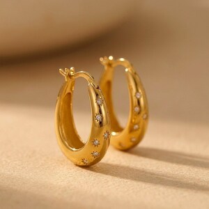 May include: A pair of gold hoop earrings featuring a polished finish and small, engraved star motifs. Each star is centered with a tiny, clear, sparkling gemstone. The hoops have a hinged closure mechanism visible at the top of the earring structure.