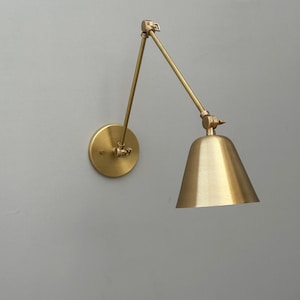 Puede incluir: Una lámpara de pared de latón con un brazo ajustable y una pantalla cónica. La lámpara tiene una base circular y un brazo giratorio para iluminación direccional. El diseño es minimalista y moderno, adecuado para varios estilos de interior. La lámpara es de latón.