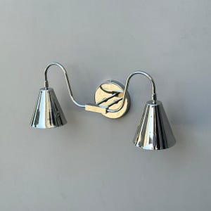 May include: A chrome wall sconce featuring two conical shades. The fixture has a curved arm design and a circular base. The shades reflect the surroundings, creating a bright, modern look. Suitable for interior lighting.