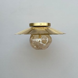 May include: A brass-colored ceiling light fixture with a unique design. It features a circular base, a wide, fluted shade, and a textured, amber-colored glass globe. The fixture is mounted on a white wall, showcasing its modern aesthetic.
