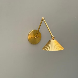 May include: A brass wall-mounted lamp with an adjustable arm and a pleated lampshade. The lamp has a circular base and a cone-shaped shade. The shade is gold in color and has a textured surface. The arm is adjustable, allowing the light to be positioned as needed.