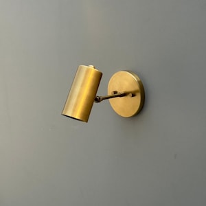 Puede incluir: Aplique de pared de latón. Tiene un cabezal cilíndrico y una base circular, conectados por un pequeño brazo. El acabado es dorado metálico. El aplique está montado en una pared gris. Sus dimensiones son de unos 10 cm de largo.