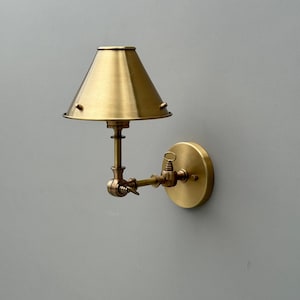 May include: A brass wall-mounted lamp with a conical shade. The lamp features an adjustable arm and a circular base. The shade is a warm gold color, and the fixture has a vintage aesthetic. The lamp is designed for indoor use.