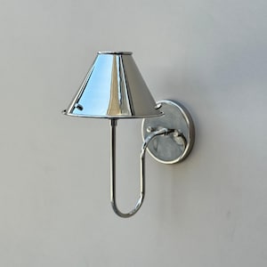 Op de afbeelding: Een gepolijste chromen wandlamp met een conische kap. De lamp heeft een cirkelvormige basis en een gebogen arm. De kap reflecteert de omgeving. De lamp is bevestigd aan een lichtgrijze muur. Dit is een moderne lamp.