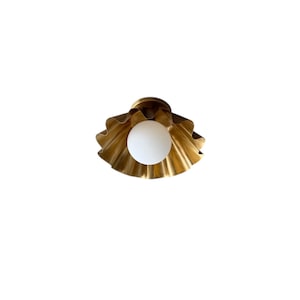 May include: A gold-colored, seashell-shaped ceiling light fixture with a white, spherical light bulb in the center. The fixture has a ruffled edge design and is mounted on a white background.