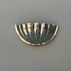 May include: A silver-coloured wall sconce with a shell-like design. The sconce has a half-circle shape with vertical, curved ridges that create a textured surface. The reflective metal surface catches the light, creating highlights and shadows.