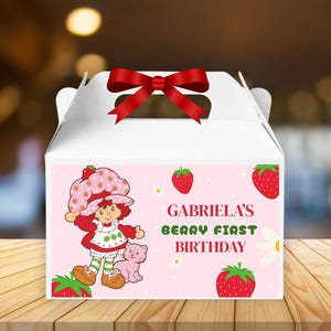 Puede incluir: Caja blanca con lazo rojo. La caja tiene un fondo rosa con un personaje de Strawberry Shortcake, fresas y el texto "Gabriela's Berry First Birthday."