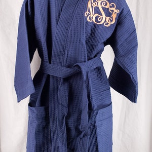 May include: A navy blue waffle weave robe with a monogram embroidered on the left chest. The robe has a self-tie belt and two pockets.