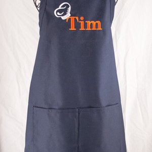 Pu&ograve; includere: Un grembiule blu navy con una grande tasca sul davanti. Il grembiule ha un cappello da cuoco bianco con la parola "CHEF" ricamata e il nome "Tim" ricamato in arancione sotto.