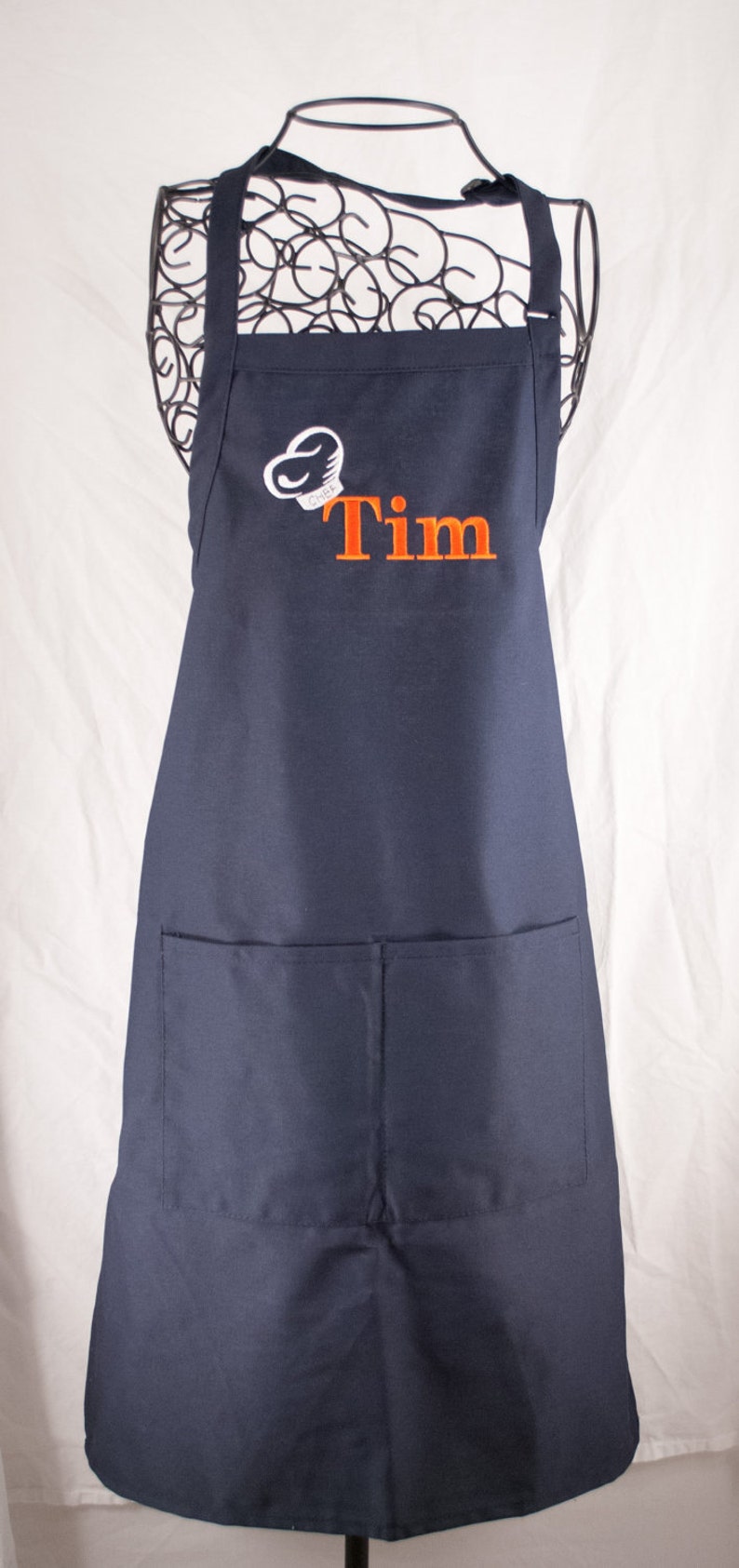 Pu&ograve; includere: Un grembiule blu navy con una grande tasca sul davanti. Il grembiule ha un cappello da cuoco bianco con la parola "CHEF" ricamata e il nome "Tim" ricamato in arancione sotto.