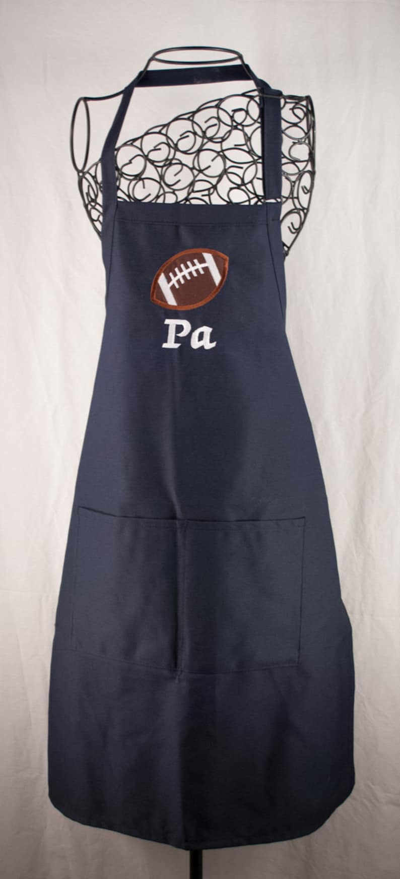 May include: A navy blue apron with a large brown football and the word "Pa" embroidered in white. The apron has a large pocket on the front.
