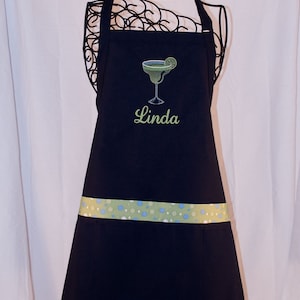 May include: A black apron with a green margarita glass and lime embroidered on the front. The apron has a green and white polka dot trim and the name "Linda" is embroidered below the glass.