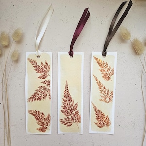 May include: Three handmade bookmarks with botanical designs. Each bookmark features a pressed fern design in shades of brown and cream. The bookmarks have cream-coloured backgrounds and are topped with satin ribbons in white, burgundy, and brown.