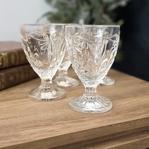 Peut inclure: Verres à pied transparents avec des motifs floraux gravés. Les verres sont disposés sur une surface en bois, avec un arrière-plan flou de verdure et de livres. Les verres sont probablement destinés à servir du vin ou d'autres boissons.