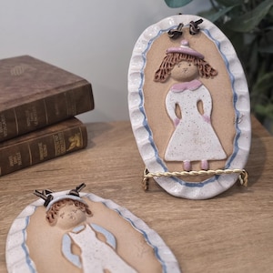 Peut inclure: Deux plaques ovales en céramique avec un motif de fille. Les plaques présentent un fond beige, une bordure blanche avec des accents bleus et un support en fil d'or. Une plaque est debout, l'autre est à plat.
