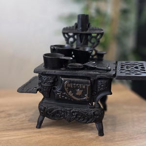 May include: A miniature black cast iron stove with several small pots and pans. The stove has the word "RESCEN" on the front and is set on a wooden surface. The stove is a decorative item.