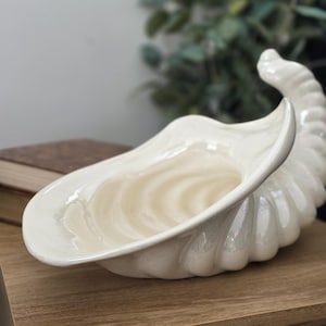 May include: A pearlescent white ceramic cornucopia-shaped decorative bowl. The bowl has a wide opening and a curved, spiraled design on the exterior. It sits on a wooden surface, with greenery in the background.