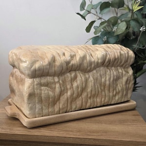 Peut inclure: Une boîte à pain en céramique en forme de pain de mie, posée sur un plateau en bois. La boîte à pain beige présente des lignes verticales, imitant la texture du pain tranché. Elle repose sur une surface en bois.