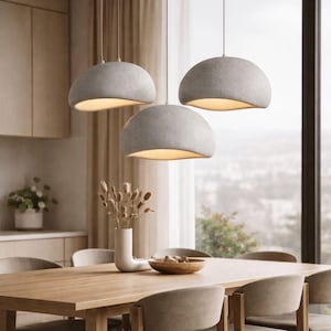 Peut inclure: Trois lampes suspendues grises à l'aspect arrondi et pierreux sont suspendues au-dessus d'une table à manger en bois. Les lampes ont une surface douce et texturée et émettent une lueur chaude. La salle à manger comprend un vase avec des plantes séchées et un bol en bois.