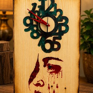 May include: A rectangular wooden wall clock with burnt edges featuring a dark teal resin clock face with raised numbers. The clock has bright red hands and a red painted graphic of a face with dripping tears on the lower half of the wood.