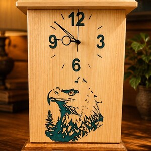 May include: A rectangular light-colored wooden clock featuring a dark teal eagle head graphic at the bottom. The clock face displays the numbers 12, 3, 6, and 9 in a dark teal font with black clock hands and a circular detail on the hour hand.