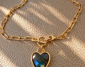 Op de afbeelding: Een goudkleurige ketting met een knevelsluiting en een hartvormige hanger. De hanger bevat een gefacetteerde labradorietsteen met blauwe en grijze iriserende flitsen, gezet in een gladde, goudkleurige metalen rand.