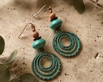 May include: A pair of drop earrings featuring a copper-toned hook, a ribbed copper bead, a small dark spacer, and a smooth turquoise-coloured bead with dark veining. The base consists of a circular, textured, oxidised teal metal spiral pendant.
