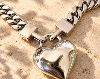 Op de afbeelding: Een zilverkleurige metalen ketting met een gepolijste, bolvormige hartvormige hanger die met twee rechthoekige metalen verbindingsstukken aan de middelste schakels is bevestigd. De ketting heeft een dikke schakelstructuur.