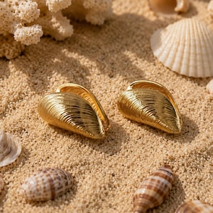 May include: A pair of gold-toned earrings shaped like textured mussel shells. The earrings feature a polished metallic finish with fine, realistic ridges mimicking the natural form of a bivalve shell, presented in a symmetrical orientation.