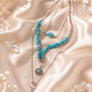Puede incluir: Un collar de varias capas en tono plateado con fragmentos de piedra color turquesa, una cuenta central ovalada color turquesa, un colgante circular de metal con patrón en relieve y centro turquesa, y un dije en forma de gota.