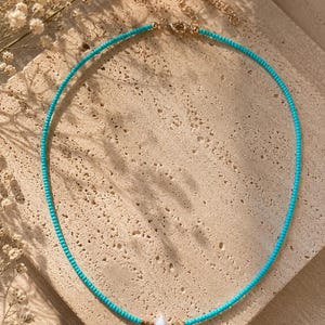 Può includere: Una collana di perline turchesi con una singola perla bianca a goccia al centro, accentuata da piccole perline color oro su entrambi i lati. La collana include una chiusura in metallo color oro e una catenella di estensione regolabile sul retro.