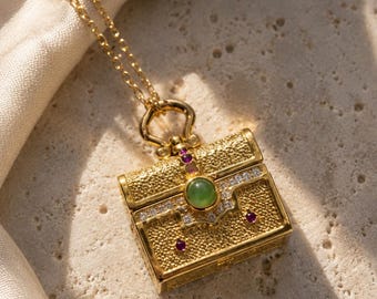 Op de afbeelding: Een goudkleurige ketting met hanger in de vorm van een vierkant, getextureerd doosje. De hanger is versierd met een centrale ronde groene steen, kleine heldere kristallen en drie kleine rode steentjes, hangend aan een fijne goudkleurige ketting.