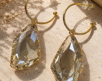 Op de afbeelding: Een paar gouden oorringen met bungelende, gefacetteerde, heldere, druppelvormige kristallen. Elk kristal is gevat in een goudkleurige metalen zetting met een getextureerde rand. De oorbellen zijn voorzien van doorzichtige plastic sluitingen.