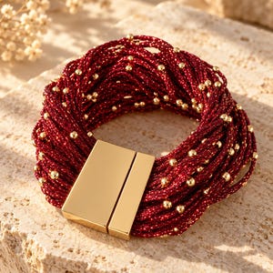 May include: A multi-strand bracelet featuring deep red, shimmering cord interwoven with small, polished gold-tone beads. The bracelet is secured with a rectangular, smooth gold-tone magnetic clasp that connects the two ends of the strands.