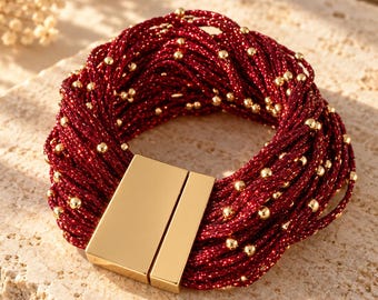 Op de afbeelding: Een meerstrengs armband bestaande uit dieprode, glinsterende koorden verweven met kleine, gepolijste goudkleurige kralen. De armband wordt gesloten met een rechthoekige, gladde goudkleurige magnetische sluiting die de uiteinden verbindt.