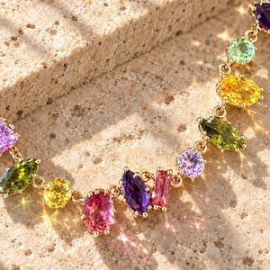 Pu&ograve; includere: Una collana a catena dorata con una serie di pietre preziose multicolori in varie forme, inclusi tagli marquise, ovali e rettangolari. Le pietre mostrano un vibrante spettro di colori come viola, rosa, giallo e verde.