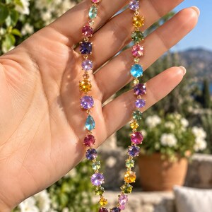 Pu&ograve; includere: Una collana a catena color oro caratterizzata da una serie di pietre preziose sfaccettate multicolori in varie forme, tra cui rotonde, ovali e a goccia. Le pietre mostrano tonalit&agrave; vivaci di rosa, blu, viola, giallo e verde, collegate in un filo continuo.