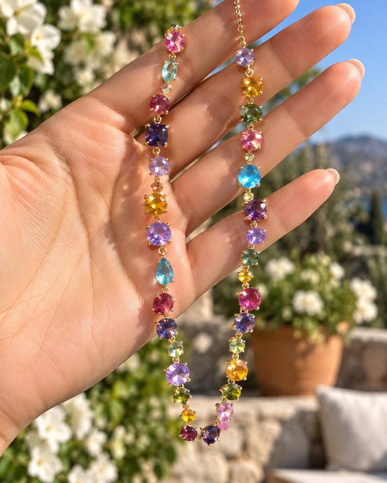 Pu&ograve; includere: Una collana a catena color oro caratterizzata da una serie di pietre preziose sfaccettate multicolori in varie forme, tra cui rotonde, ovali e a goccia. Le pietre mostrano tonalit&agrave; vivaci di rosa, blu, viola, giallo e verde, collegate in un filo continuo.