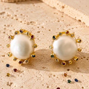 May include: A pair of gold stud earrings featuring a large, round, white pearl at the center. The pearl is surrounded by a gold frame set with small, multi-colored gemstones including red, blue, green, and yellow stones, with loose stones nearby.