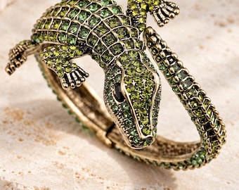 Op de afbeelding: Een scharnierende armband in de vorm van een krokodil, gemaakt van antiek goudkleurig metaal. Het oppervlak is ingelegd met talloze kleine, gefacetteerde groene kristallen in diverse tinten, voorzien van zwarte stenen ogen en een geschubde textuur.