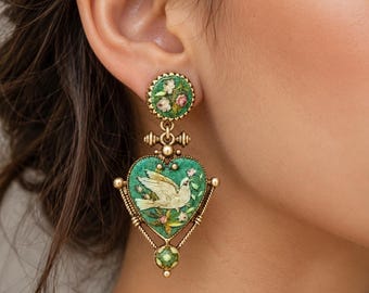 Op de afbeelding: Een goudkleurige hangende oorbel met een ronde knop met bloemmotief en een hartvormige hanger. De hanger toont een illustratie van een witte duif op een getextureerde groene achtergrond, geaccentueerd met kleine bloemdetails en een kleine groene kraal.