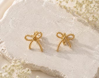 May include: A pair of gold-toned stud earrings shaped like bows. Each earring features a textured, beaded surface that forms the loops and tails of the bow design. The earrings are metallic with a polished finish, showcasing a symmetrical structure.