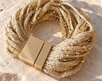 Op de afbeelding: Een goudkleurige armband bestaande uit meerdere gevlochten metalen strengen verweven met kleine gouden kralen. De armband is voorzien van een rechthoekige, gladde goudkleurige magnetische sluiting die de uiteinden van de strengen bij elkaar houdt.