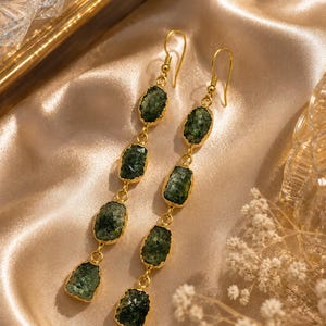 May include: A pair of long drop earrings featuring four vertically aligned, rough-cut dark green stones set in gold-toned metal bezels. Each earring hangs from a simple gold-toned hook, showcasing a textured, natural appearance for each stone.