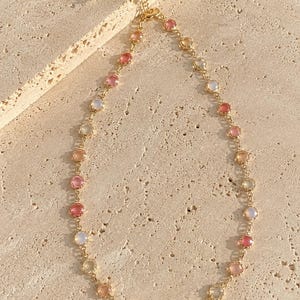 Può includere: Una collana a catena color oro caratterizzata da una serie di pietre rotonde incastonate in varie tonalità di rosa, pesca e bianco traslucido. La collana include una delicata catena a maglie con chiusura a moschettone e catenella regolabile.