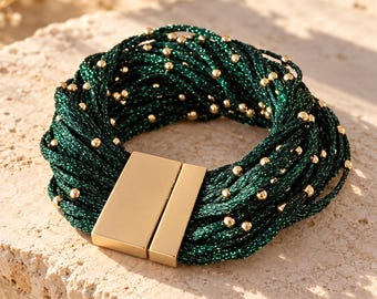 Op de afbeelding: Een meerstrengs armband bestaande uit talloze donkergroene metallic draden die met elkaar zijn verweven, geaccentueerd met kleine goudkleurige kraaltjes. De armband wordt gesloten met een rechthoekige, gepolijste goudkleurige magneetsluiting.
