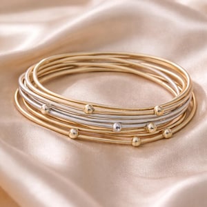 May include: A stack of gold and silver bangles with small, round, gold-colored beads. The bangles are arranged on a light peach-colored satin fabric, creating a luxurious and elegant aesthetic. The jewelry is simple and classic.