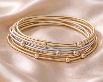 Op de afbeelding: Een stapel dunne, ronde armbanden in afwisselende goud- en zilverkleurige tinten. Elke individuele armband is voorzien van een kleine, gladde, bolvormige kraal die in de metalen draad is geïntegreerd voor een verfijnde, gelaagde uitstraling.